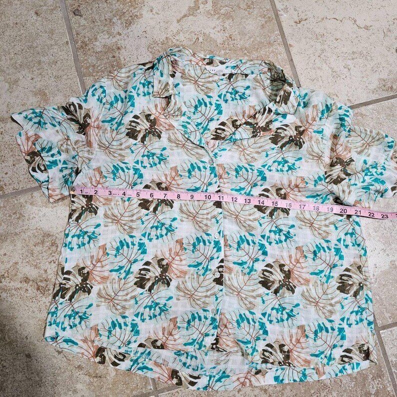 May include: A short-sleeved button-down shirt with a tropical leaf pattern in shades of brown, teal, and pink on a white background. The shirt is measured at approximately 23 inches across the chest.