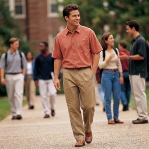 May include: A short-sleeved button-down shirt in a muted coral color with a single chest pocket, paired with khaki-colored pleated trousers, a brown leather belt, and brown leather loafers, worn by a person walking outdoors.