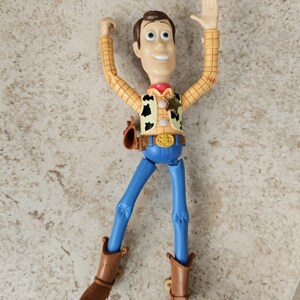 May include: A Woody action figure from the Toy Story franchise. The figure has a tan plaid shirt, blue pants, brown boots, and a sheriff's badge. The figure's arms are raised, and it has a friendly expression.