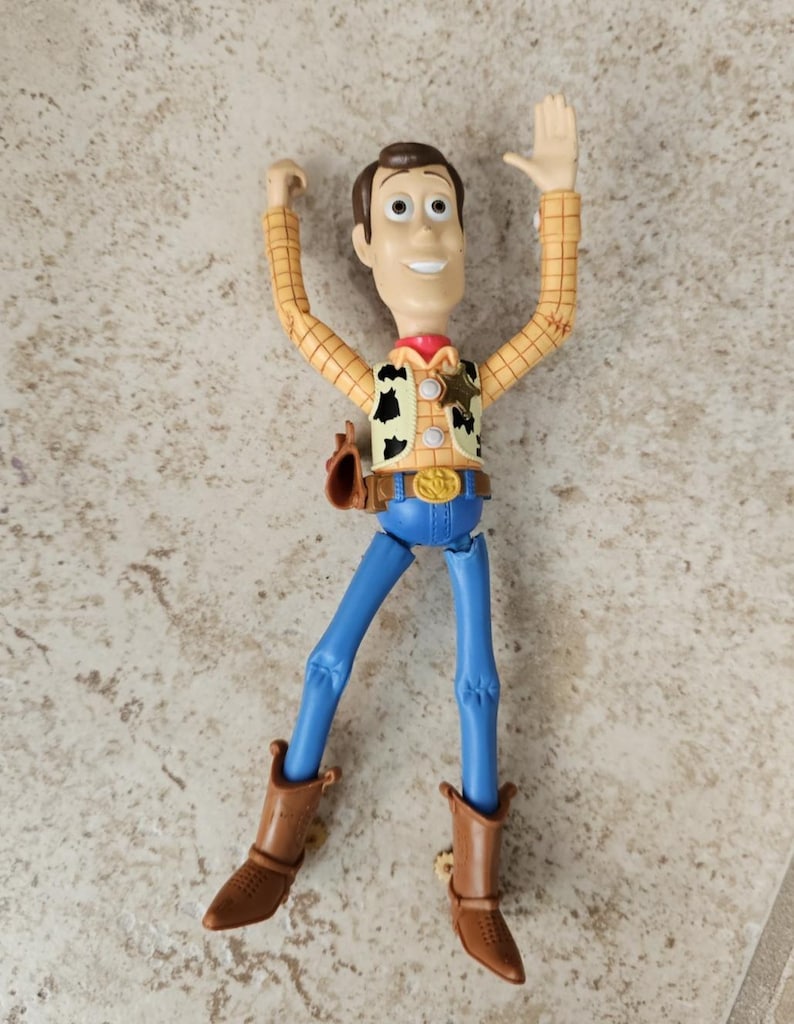 May include: A Woody action figure from the Toy Story franchise. The figure has a tan plaid shirt, blue pants, brown boots, and a sheriff's badge. The figure's arms are raised, and it has a friendly expression.