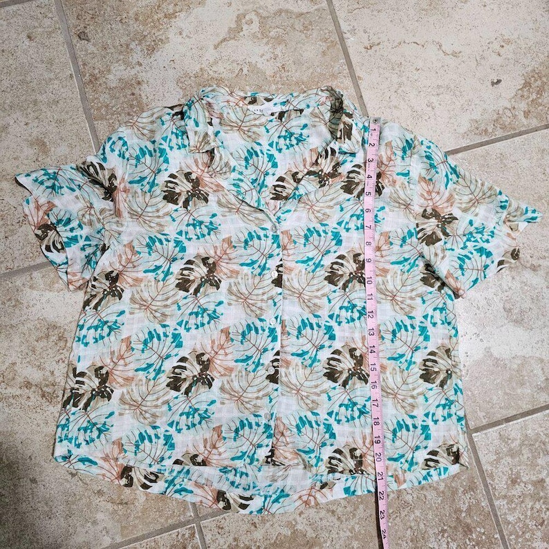 May include: White button-down shirt with a tropical leaf pattern in teal, brown, and tan. The shirt has short sleeves and a collared neckline. The length of the shirt is approximately 23 inches.
