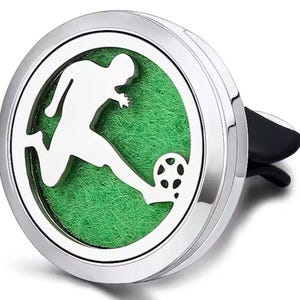 May include: A silver car air freshener featuring a white silhouette of a footballer kicking a ball, set against a green background. The freshener has a clip for attachment.