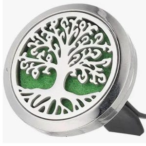 May include: A silver car air freshener with a tree of life design. The circular freshener has a detailed cutout of a tree against a green background. A black clip is attached for easy placement in a car's air vent.