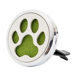 Peut inclure: Un diffuseur de parfum pour voiture argenté avec un motif de patte de chien. La patte est découpée dans le métal argenté, révélant un intérieur vert texturé. Une pince noire est fixée au dos pour l'accrocher.