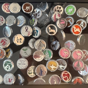 May include: A selection of round, silver-coloured snap jewellery charms with diverse designs, including animals, symbols, and text. The charms are displayed in a black tray, showcasing a variety of colours and patterns. Some charms feature images of dogs, flamingos, and hearts.