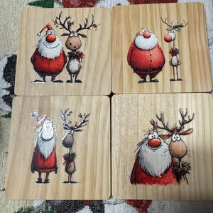 May include: Four square wooden coasters with festive Christmas designs. Each coaster features a Santa Claus and reindeer illustration. The Santa figures are in red suits with white beards, and the reindeer have antlers and decorations.