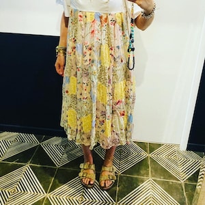 Peut inclure: Une robe blanche avec une jupe à imprimé floral patchwork et un corsage en dentelle blanche. La robe a des manches courtes et un décolleté carré. Le mannequin porte des sandales marrons.