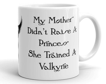 May include: White ceramic coffee mug with black text that reads "My Mother Didn't Raise A Prince... She Trained A Valkyrie".