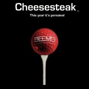May include: A red golf ball with the word "DEEMO" printed on it, sitting on a white golf tee against a black background. The text "Revenge of the Cheesesteak" is written in white above the golf ball, and the text "This year it's personal" is written in white below the golf ball. The text "Boom Boom goes the Dynamite!" is written in white at the bottom of the image, and the text "D. Monosson" is written in red below that.