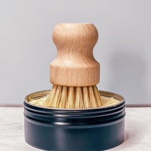 Op de afbeelding: Een zwarte metalen blik met een borstel met houten handvat erin. Het blik is gevuld met een lichtgele substantie.