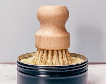 Puede incluir: Una lata de metal negra con un cepillo de mango de madera en su interior. La lata está llena de una sustancia de color amarillo claro.