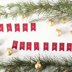 Könnte beinhalten: Rote Filzbanner mit weißen Buchstaben, die "Frohe Weihnachten!" buchstabieren, hängen an einem weißen Hintergrund mit goldenen Kugeln und Kiefernästen.