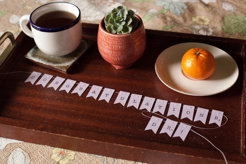 May include: A wooden tray with a cup of tea, a small potted succulent plant, a plate with an orange, and a banner that reads "Congratulations Dr."