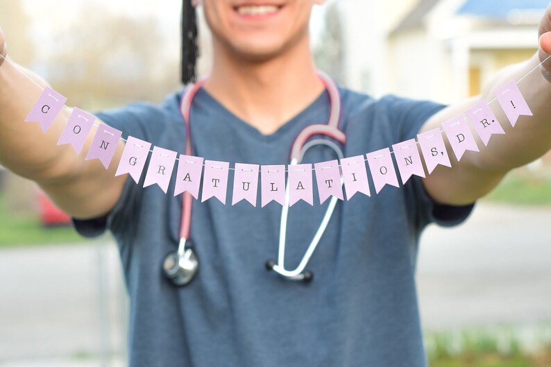 May include: A person wearing a blue scrubs top and a stethoscope holds up a pink banner that reads "Congratulations, Dr.!".