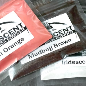 May include: Two small plastic bags containing mica powder. The first bag contains a bright orange powder with the label "Iridescent Mica Powder - Peach Orange". The second bag contains a dark brown powder with the label "Iridescent Mica Powder - Mudbug Brown".