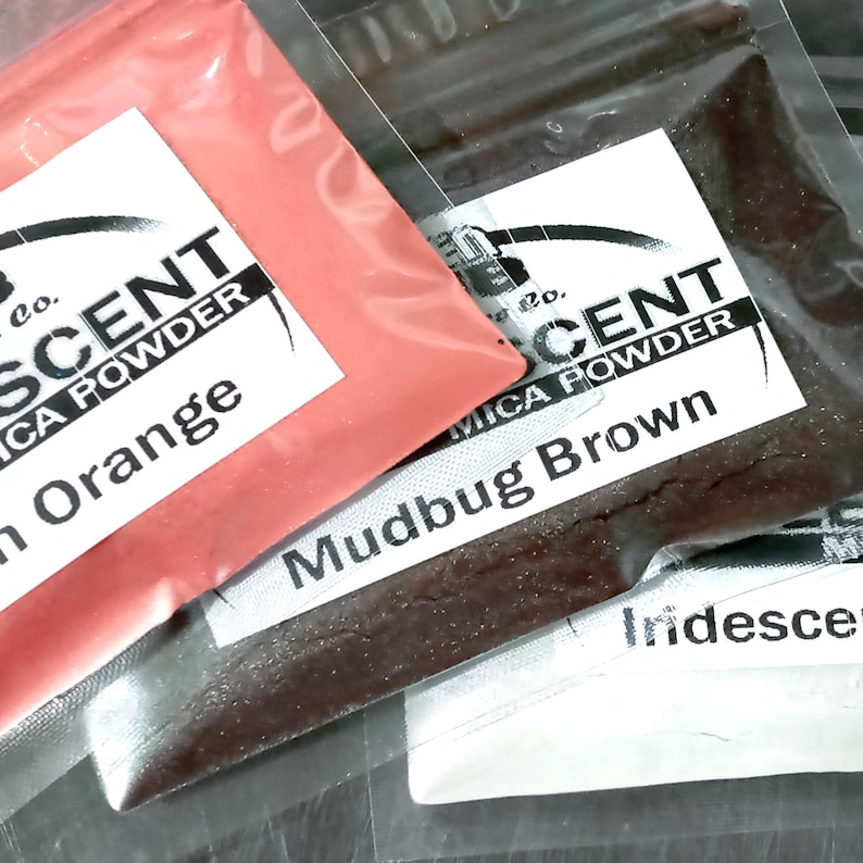 May include: Two small plastic bags containing mica powder. The first bag contains a bright orange powder with the label "Iridescent Mica Powder - Peach Orange". The second bag contains a dark brown powder with the label "Iridescent Mica Powder - Mudbug Brown".