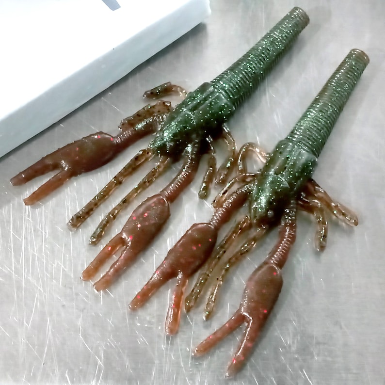 May include: Two green and brown crawfish-shaped fishing lures with multiple legs. The lures have a textured, glittery finish.