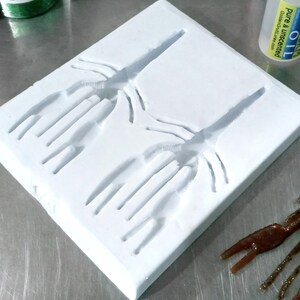 May include: A white silicone mould with two impressions of a crawfish or crayfish. The mould is on a silver surface.