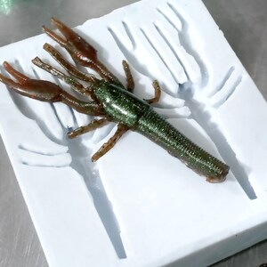 May include: A white silicone mould for making a green and brown crawfish lure. The mould has a detailed design of a crawfish with claws and a body.