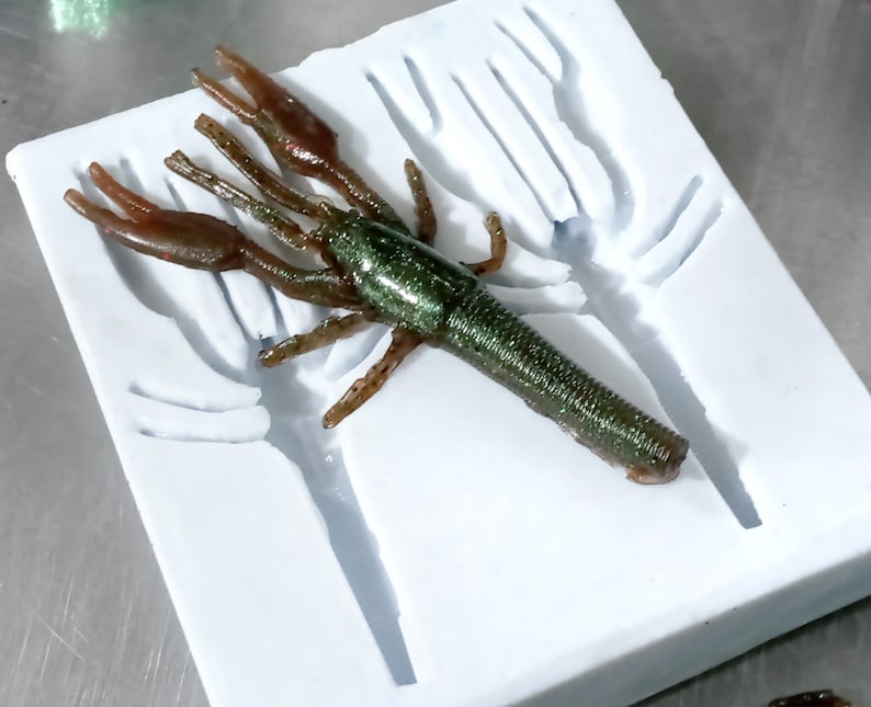 May include: A white silicone mould for making a green and brown crawfish lure. The mould has a detailed design of a crawfish with claws and a body.