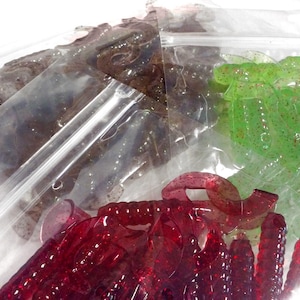 May include: Assortment of plastic fishing lures in clear plastic bags. The lures are in various colors, including red, green, and brown.
