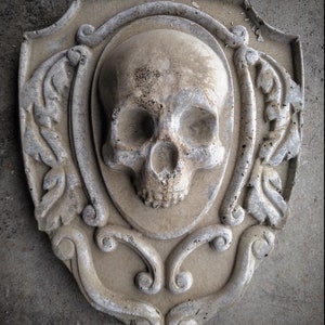 May include: A gray concrete plaque with a detailed, ornate design featuring a sculpted human skull in the center.