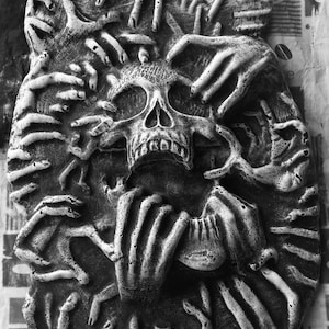 May include: A black and white sculpture of a skull surrounded by many hands. The sculpture is made of a rough, textured material.
