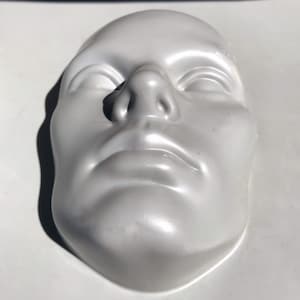 May include: A white plaster cast of a human face. The face is expressionless and the eyes are closed.