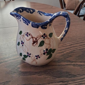 May include: A ceramic pitcher with a cream-colored base, adorned with blue trim and a handle. The pitcher features a floral design with brown vines, green leaves, and small blue and purple flowers. The pitcher is on a wooden table.