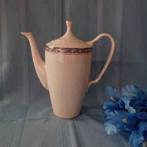 May include: A white porcelain coffee pot with a lid and a curved spout. The pot has a gold trim and a decorative band with a blue and gold pattern. The handle is curved and the pot is on a light blue surface. A blue flower is in the background.