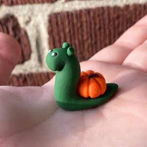 Puede incluir: Un caracol de arcilla verde con una pequeña calabaza naranja en la espalda.