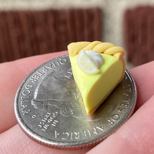 Puede incluir: Una rebanada de pastel de limón merengue en miniatura hecha de arcilla polimérica, junto a una moneda de 25 céntimos de euro para la escala. El pastel es amarillo con una cobertura de merengue blanco y una rodaja de limón en la parte superior.