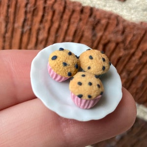 May include: Three miniature chocolate chip muffins on a white plate. The muffins are brown with dark chocolate chips and pink paper liners.