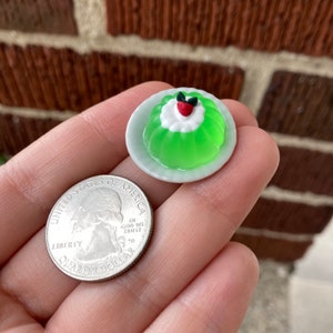 Könnte beinhalten: Ein Miniatur-Dessert aus grüner Limetten-Gelee mit einem roten und weißen Klecks Schlagsahne und einer grünen Kirsche obenauf, auf einem weißen Teller. Das Dessert wird in einer Hand neben einer US-amerikanischen 25-Cent-Münze gehalten.