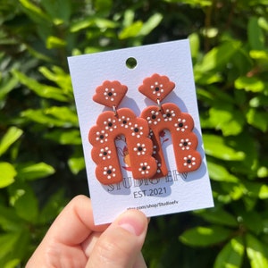 May include: Terracotta-colored polymer clay earrings with a floral design. The earrings have a scalloped arch shape with white and black dot details. The earrings are on a white card with the text "STUDIO EFV EST.2021".