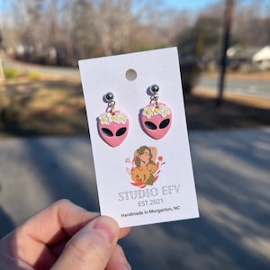 May include: A pair of pink alien head earrings with black eyes and a white daisy crown. The earrings are hanging from silver posts. The earrings are displayed on a white card with the text "STUDIO EFV EST.2021 Handmade in Morganton, NC".