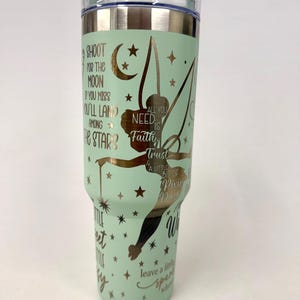 May include: A mint green stainless steel tumbler with a clear lid and straw. The exterior features a metallic gold fairy silhouette, stars, and crescent moon. Text includes: Shoot for the moon if you miss you'll land among the stars, All you need is faith, trust & a little bit of pixie dust, and leave a little sparkle wherever you go.