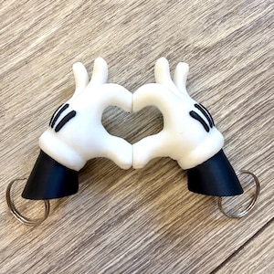 May include: A pair of white cartoon-style glove keychains with black wrist cuffs, each featuring three black lines on the back of the hand. The two glove figures are positioned to form a heart shape with their fingers, and each has a metal ring attached.