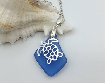 May include: A blue sea glass pendant necklace with a silver turtle charm. The necklace is displayed on a white seashell.