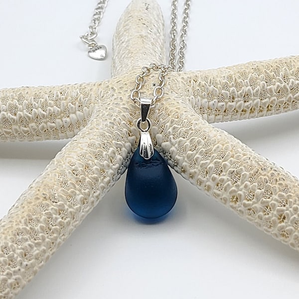 Könnte beinhalten: Eine silberne Kettenkette mit einem dunkelblauen Glas-Tropfenanhänger. Die Kette wird auf einem weißen Seestern präsentiert.