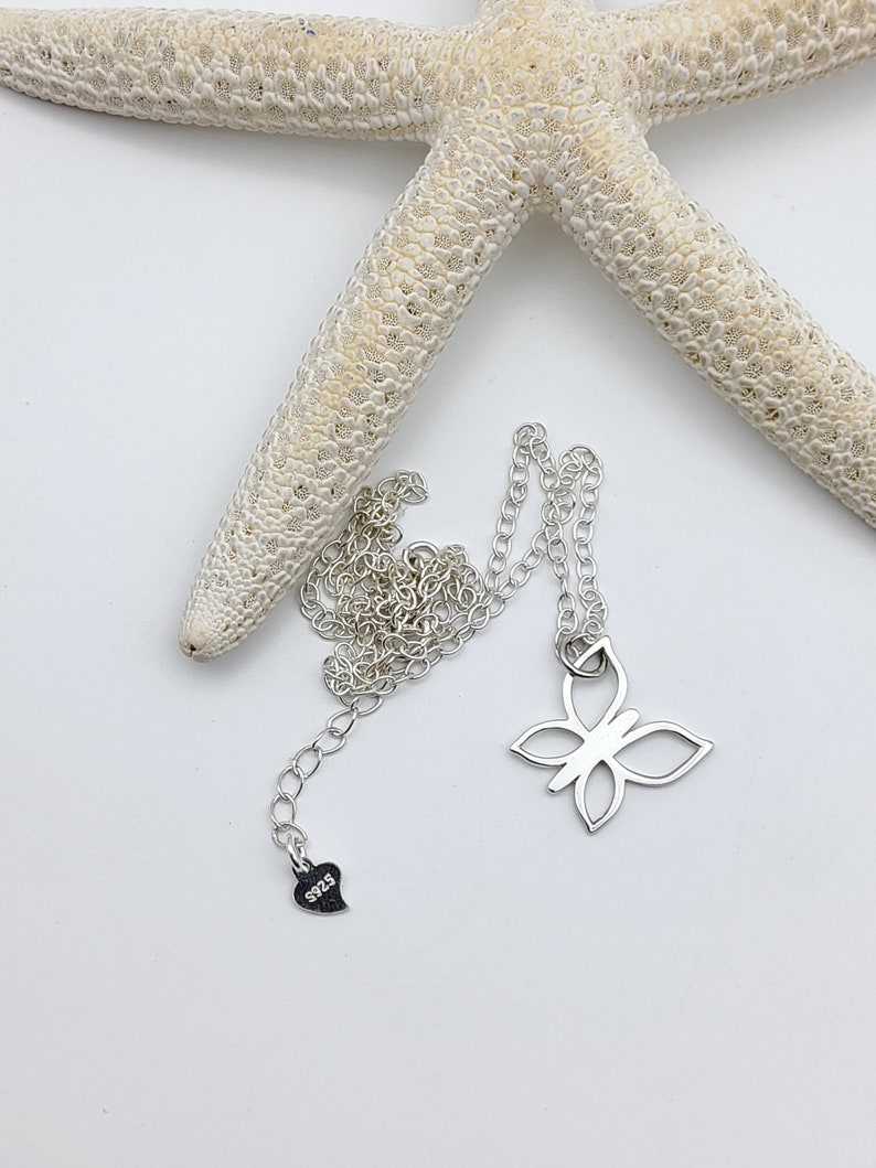 May include: A silver butterfly necklace with a heart charm that says "925". The necklace is on a white background with a starfish.