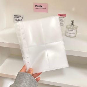 May include: Clear, multi-pocketed binder sheet with a row of holes along the left edge. The sheet is held in front of a white shelf with a perfume bottle and pink sticky notes. Text on the sticky notes reads "Pink." and "BECAUSE I COUL--D NOT STOP FOR DEA."