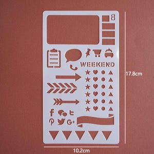 May include: A white plastic stencil with various shapes and icons for journaling or crafting. Includes shapes like arrows, stars, hearts, speech bubbles, and social media logos. The stencil is 17.8cm (7 inches) tall and 10.2cm (4 inches) wide, with the word "WEEKEND" printed on it.