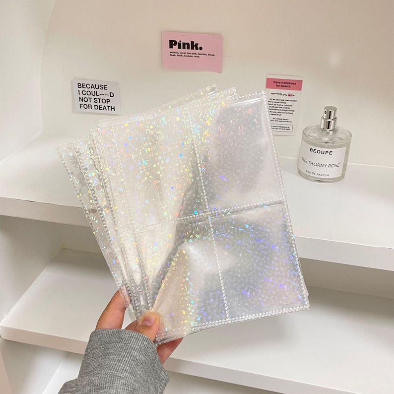 May include: Clear, sparkly photo album pages with multiple pockets. The pages are held by a hand, and the background includes a shelf with a perfume bottle and pink sticky notes that read "Pink" and "BECAUSE I COUL--D NOT STOP FOR DEATH."