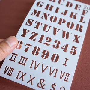 May include: A white plastic stencil featuring uppercase letters, numbers, and Roman numerals in a classic font. The stencil is held against a brown background, with the cut-out letters and numbers visible.