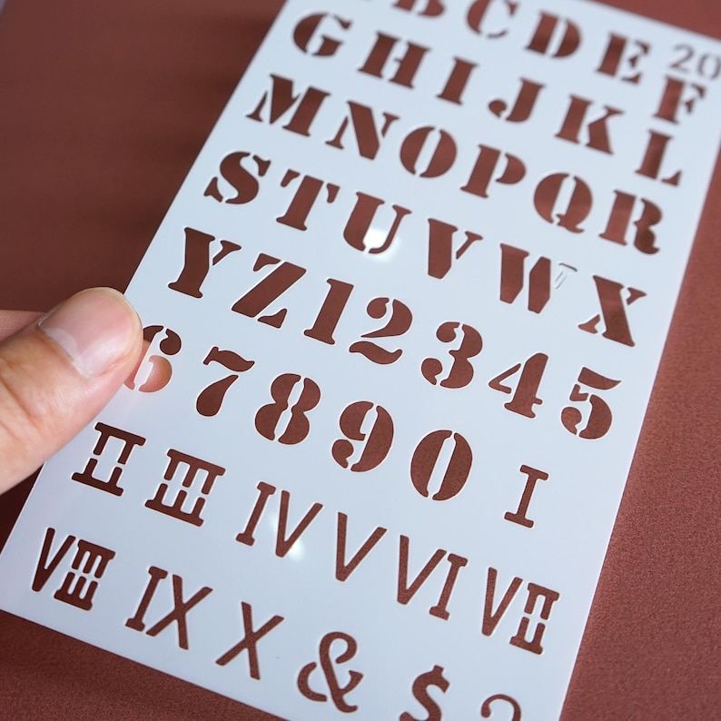 May include: A white plastic stencil featuring uppercase letters, numbers, and Roman numerals in a classic font. The stencil is held against a brown background, with the cut-out letters and numbers visible.