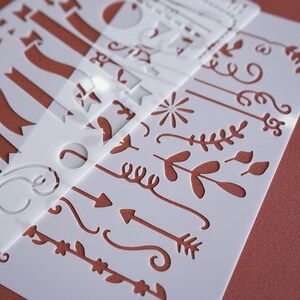 May include: A collection of white plastic stencils featuring various decorative designs. The stencils include ribbons, arrows, floral elements, and other ornamental shapes. The background is a solid reddish-brown color.