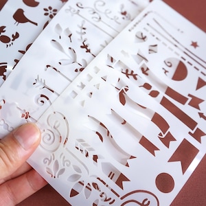 May include: A collection of white plastic stencils featuring various designs. The stencils include images of birds, flowers, arrows, banners, and geometric shapes. The stencils are held in a hand, showcasing the variety of patterns available for crafting and art projects.