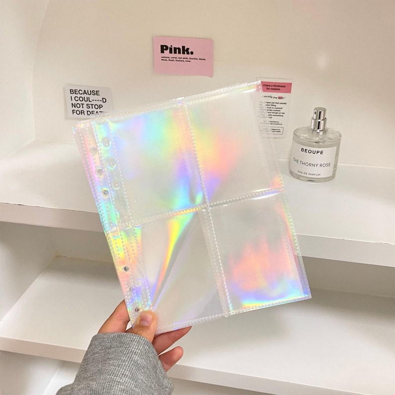 May include: A clear, iridescent photo album page with four pockets. The page is held up, displaying a rainbow sheen. The album page has multiple holes for binding. The background includes a perfume bottle and a pink sticky note that says "Pink."