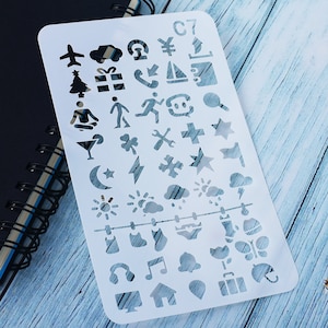 May include: A white plastic stencil featuring cut-out shapes like an aeroplane, a Christmas tree, a gift box, and a musical note. The stencil also has the text "C7". It is placed on a wooden surface. The stencil is for crafts.